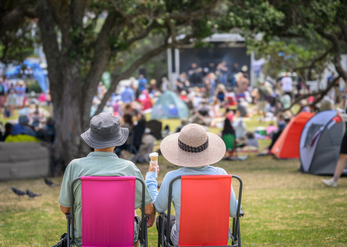 Couple sitting on camping chairs, unrecognizable crowds listen to the music concert in the park